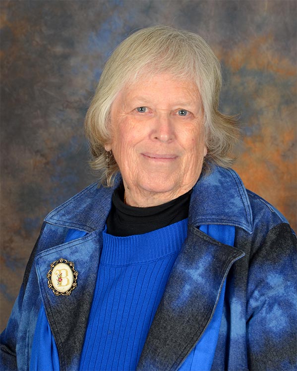 Dr Kathleen Bishop is looking at the camera with a closed mouth smile. She has light blond and brown hair, blue eyes and is wearing a blue seater over a black top. She has a blue and dark gray jacket over top with a pin on the right lapel with the letter “B”.