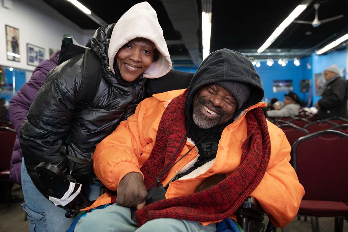 Samika Frazier and Tony Phillips are side by side and smiling at the camera. Samika is wearing a black winter puffy coat with a cream sweatshirt underneath. Her sweatshirt hood is pulled up over her baseball cap. Tony is wearing an orange winter coat with a black sweatshirt underneath. His sweatshirt hood is pulled up over his dark gray winter hat. He has on a red and black winter scarf on. Tony is a wheelchair user. They are inside in a conference room.