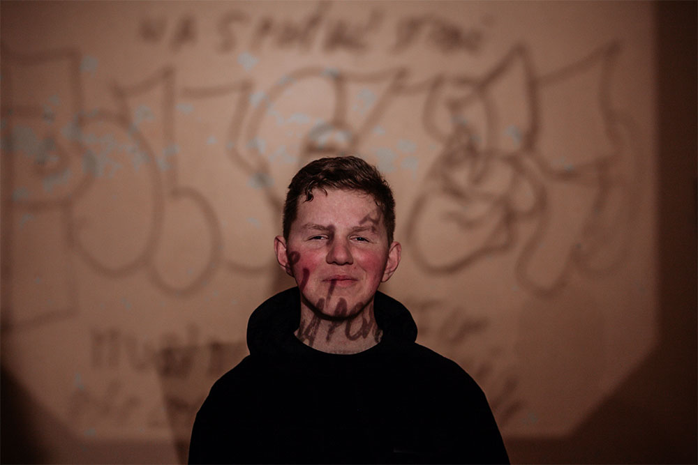 A young artist stands in front of a projected image. Handwritten words, scrawled in black marker, appear on the screen behind him and across his face. He wears a black turtleneck sweater and smiles into the camera.