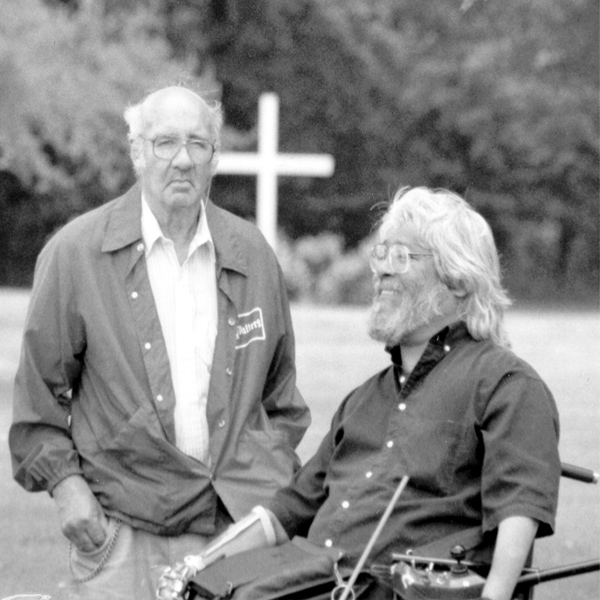 Hombre mayor de pie junto a un hombre en una silla de ruedas eléctrica con una gran cruz blanca al fondo.