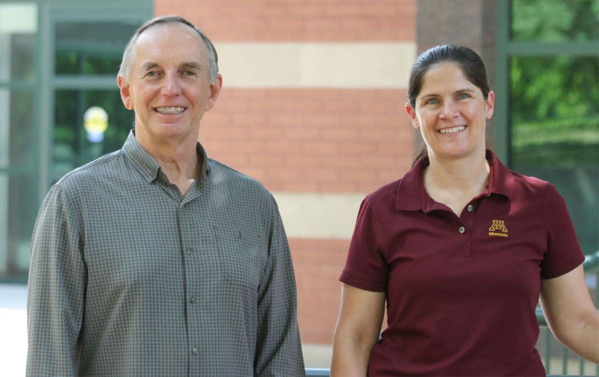 Roger and Sarah are standing outside in front of the Masonic Institute on the Developing Brain at the University of Minnesota. Roger is on the left. He is smiling and looking at the camera. He has short gray hair and is wearing a long sleeve, button down black and gray shirt. Sarah in on the left, she is smiling and looking at the camera, She has long dark brown hair that is pulled back into a ponytail and is wearing a short sleeve University of Minnesota polo shirt.
