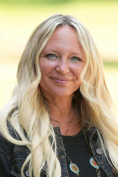 A woman with light skin tone and long, wavy blonde hair is smiling. She is wearing a black top, a dark denim jacket, and a necklace with turquoise and orange circular pendants. The background is softly blurred, featuring greenery.