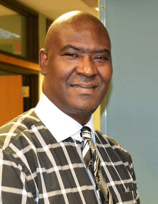 A bald man smiles as he looks straight ahead, wearing a collared, checked shirt and tie.