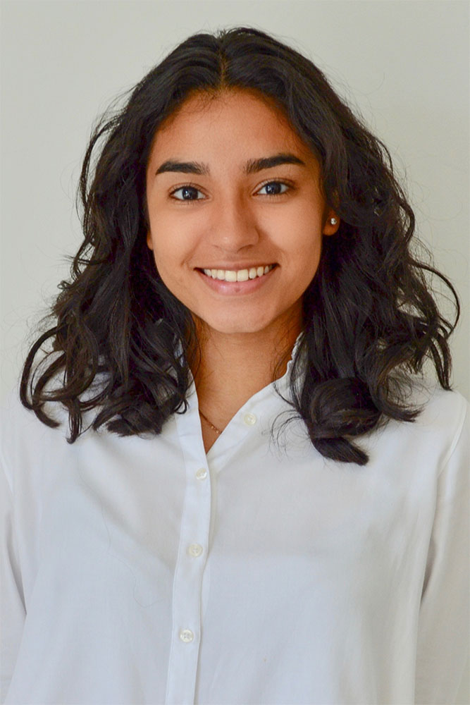 Retrato de una persona con cabello oscuro y rizado hasta los hombros, vestida con una camisa blanca abotonada. El fondo es liso y de color claro, lo que crea una apariencia limpia y profesional.