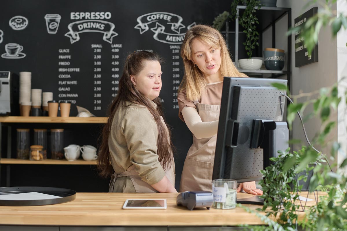 Dos mujeres trabajando en una cafetería detrás del mostrador. La mujer de la derecha está señalando algo en la pantalla de la computadora, explicándole algo a la mujer (Beth) de la izquierda. La mujer de la izquierda tiene síndrome de Down.