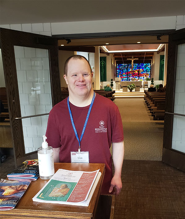 Tim is standing outside of the sanctuary at this church. In the background there is a large stained-glass window, a cross hanging in front of the it, and a pulpit. Tim is standing by the ushers stand that has church bulletins, hand sanitizer, a magazine and monthly devotional books. Tim is smiling. He has short dark brown hair, is wearing a maroon t-shirt with a church logo on the left front and a blue lanyard with a name holder with his name.