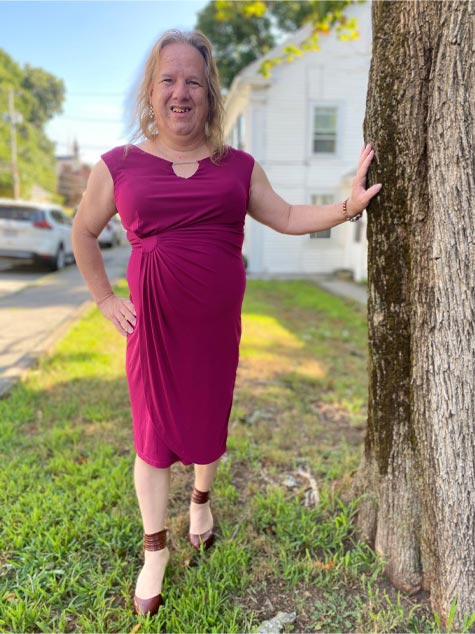 Una mujer transexual con el cabello largo hasta los hombros y vestida de magenta está de pie con una mano en la cadera y la otra apoyada en el tronco de un árbol.