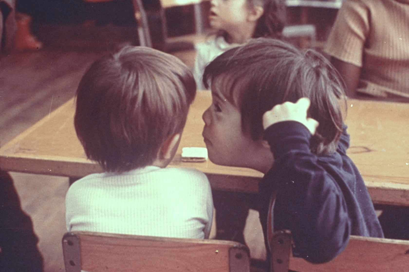 In a faded photo from the 1970s, a young school child with a disability speaks to a student without a disability. The children, who are approximately six years old, sit together in an integrated classroom.