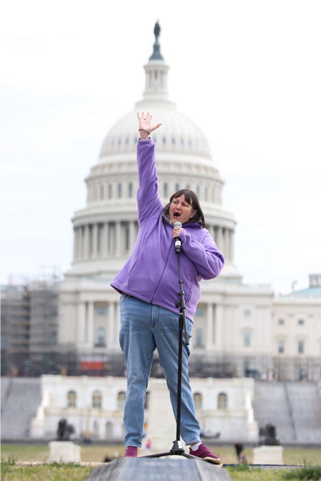 A woman wearing a purple shirt and jeans stands with one hand raised high as she speaks into a microphone while standing in front of the U.S. Capitol.