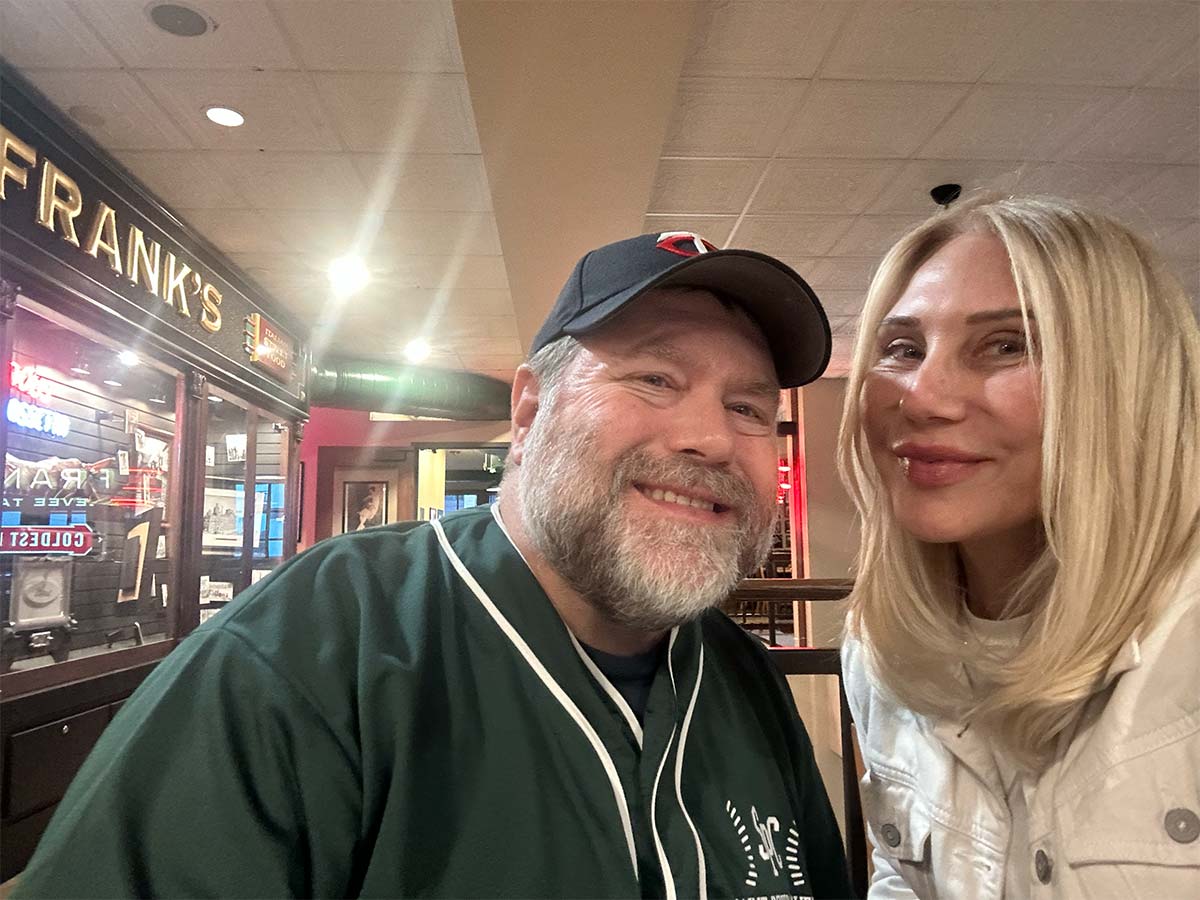 Two people standing by each other. Jimmy is the man on the left has short gray and white hair and beard. He is smiling, He is wearing a Minnesota Twins baseball hat and wearing a green and white baseball jersey. Maris is Jim’s sister and is the woman on the right, is smiling, has shoulder length blonde hair and is waring a white t-shirt and cream color denim jacket.