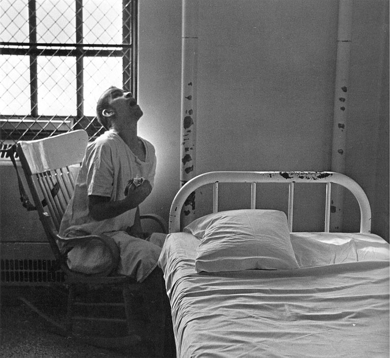 An institutionalized patient gazes at ceiling from a chair that sits next to a bed.