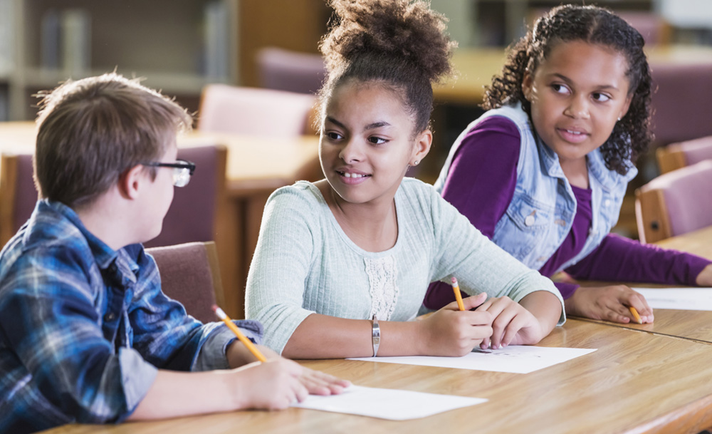 A group of 3 students, one boy, and two girls are sitting at a table together. They each hold a pencil, and each student has a piece of paper in front of them. They appear to be working on an assignment together.