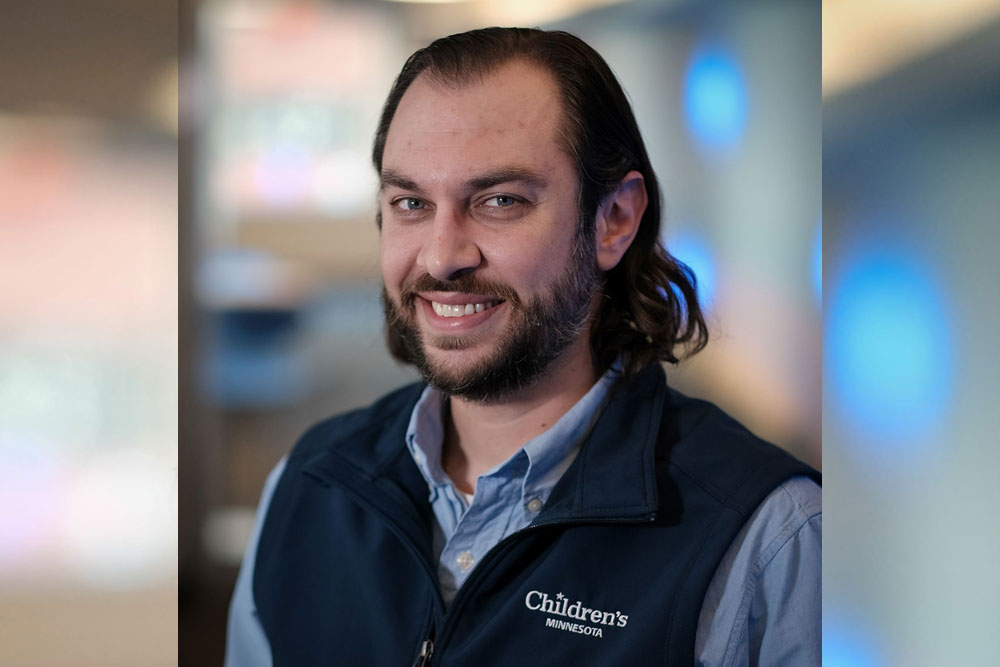 Adam Langenfeld. He is smiling at the camera and wearing a vest with the Children's Minnesota hospital logo embroidered on it. The background is blurred.