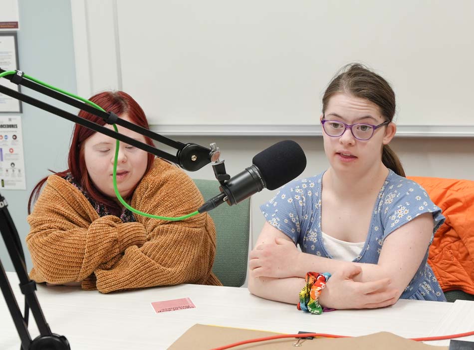 Una mujer adulta joven con gafas y una pulsera de colores habla por un micrófono mientras está sentada junto a otra mujer adulta joven con los brazos cruzados sobre una mesa.