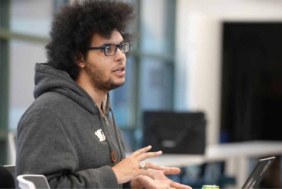 A man with a large, natural afro, wearing a hooded sweatshirt and dark-rimmed glasses, gestures as he speaks.