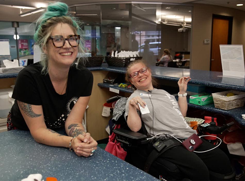 Two women wearing glasses in an office setting smile at the camera. One has blue-green hair in a messy bun and tattoos and the other has her hair pulled back and sits in a wheelchair.
