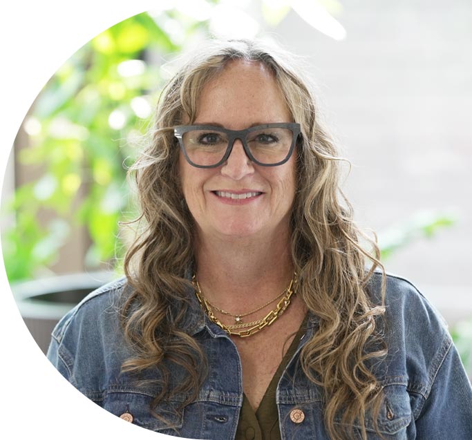 A woman with long sandy blonde, curly hair, glasses, and a jean jacket, smiling.