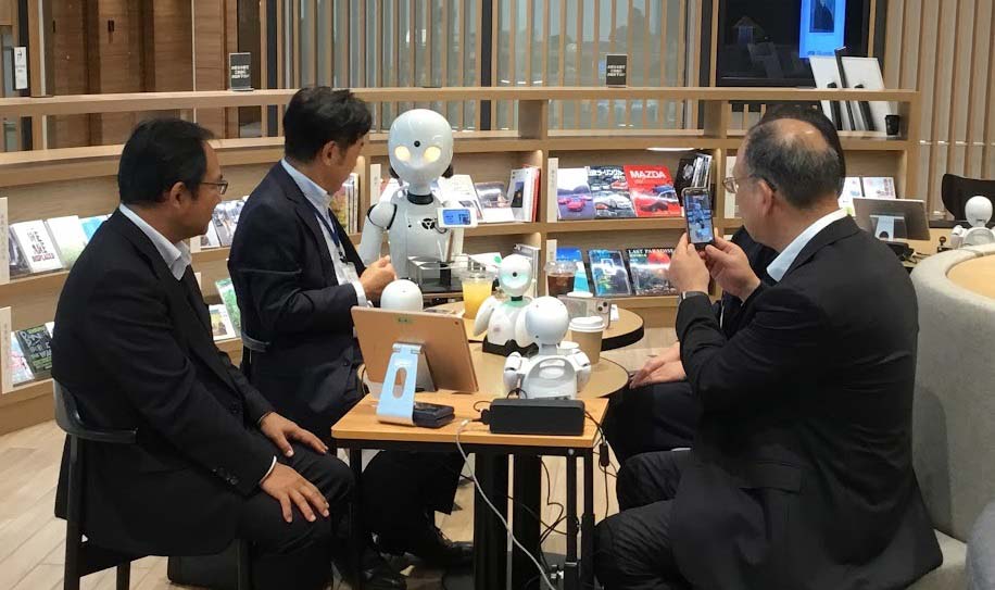 A group of four men wearing dark business suits sit at a table, looking at a robot.