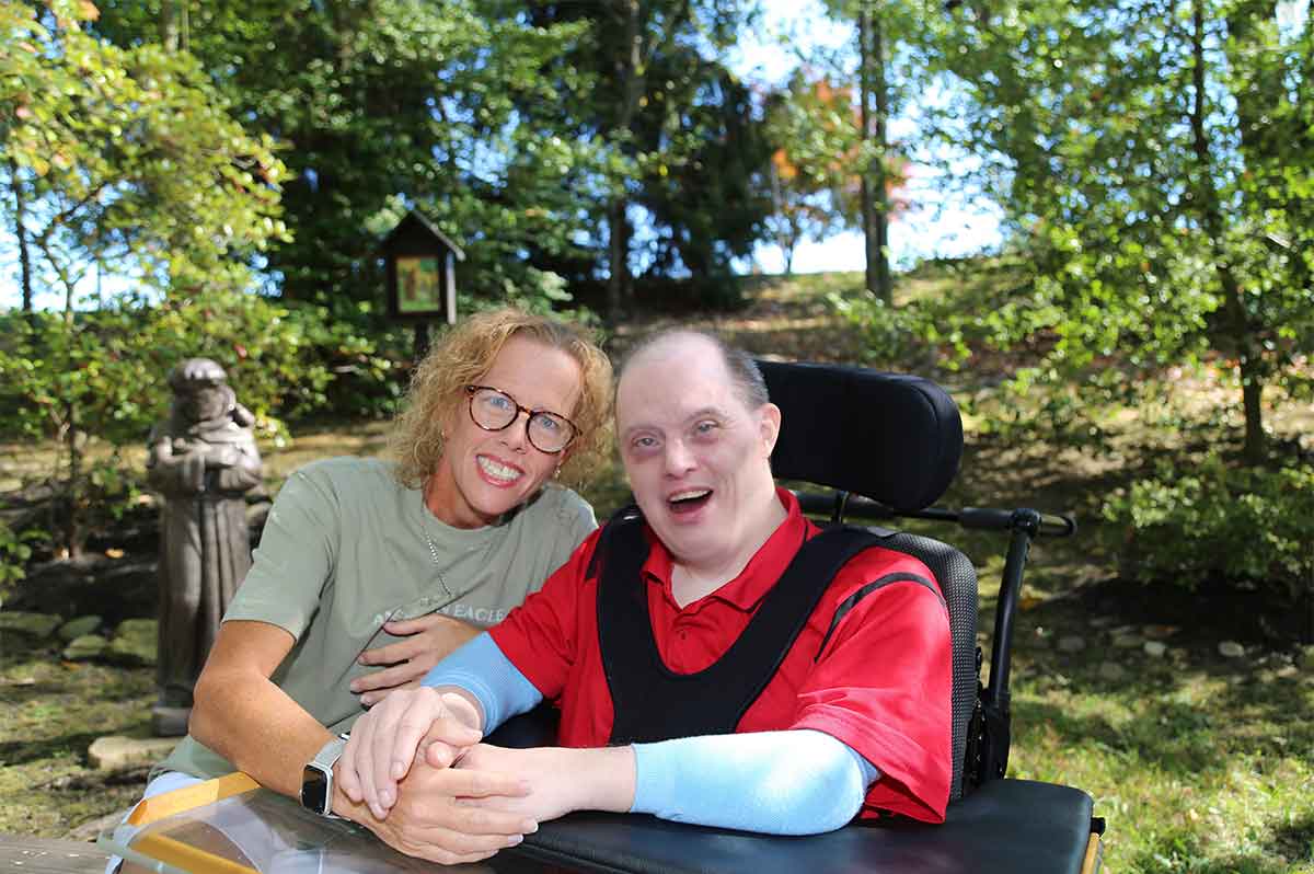 Michael y Crysal están fuera, en un parque. Ambas miran a la cámara. Crystal está a la izquierda, sentada en un banco. Su cuerpo se inclina hacia Michael. Su brazo izquierdo descansa sobre la bandeja de su regazo. Ella le está sujetando las manos. Tiene el cabello castaño hasta los hombros, lleva gafas y viste una camiseta verde musgo de American Eagle y pantalones cortos blancos. Michael sonríe y lleva una camisa polo roja con un cubrebrazos azul claro sobre los aparatos ortopédicos de los brazos y unos pantalones cortos de color beige. Tiene el cabello corto y oscuro, con entradas. Tiene ambos brazos apoyados en la bandeja del regazo y sostiene la mano de Crystal. Es usuario de silla de ruedas.