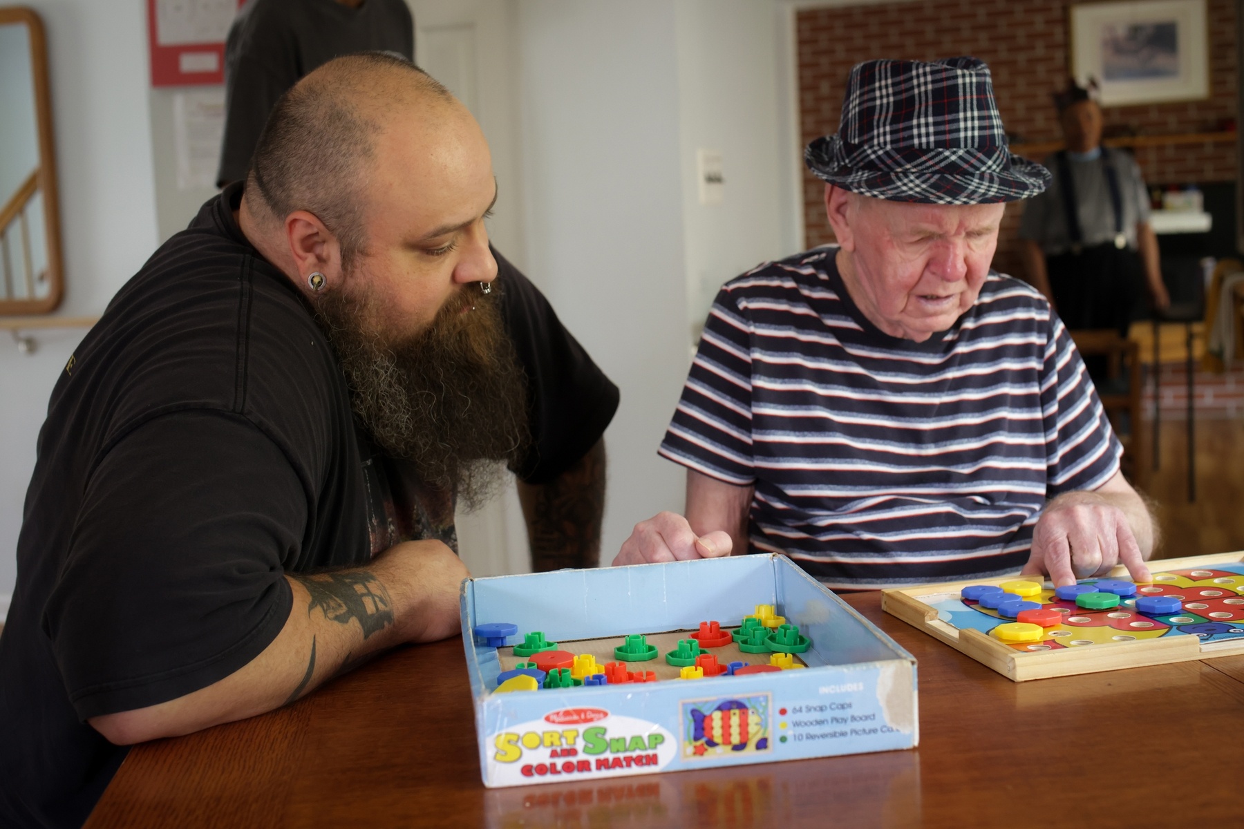 Dos hombres sentados a la mesa del comedor jugando a un juego llamado Sort and Snap, Color match. Tom es el hombre de la izquierda. Tiene el cabello corto y castaño oscuro, con una gran barba castaña oscura y blanca, un piercing en la nariz, un pendiente, un tatuaje en el antebrazo derecho y lleva una camiseta negra. Wayne es el hombre de la derecha. Lleva un sombrero a cuadros negros, blancos y rojos y una camisa a rayas negras, blancas, grises y rojas. Está mirando hacia abajo y tiene la mano sobre el tablero del juego.