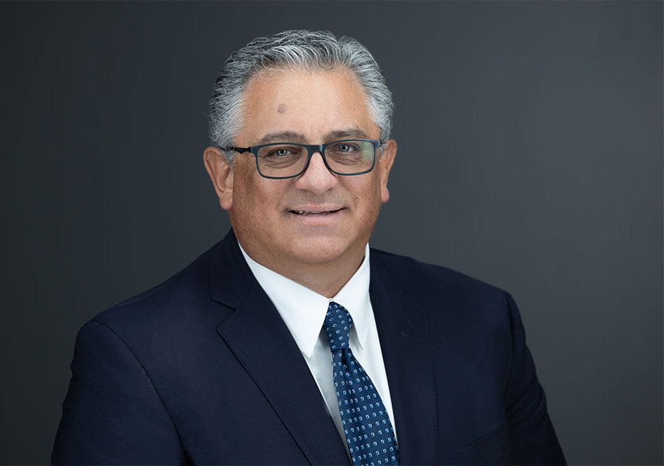 Man wearing a dark suit, white oxford style shirt with a blue and white tie, is smiling slightly, looking at the camera. He is wearing dark framed glasses and has silver and black hair.