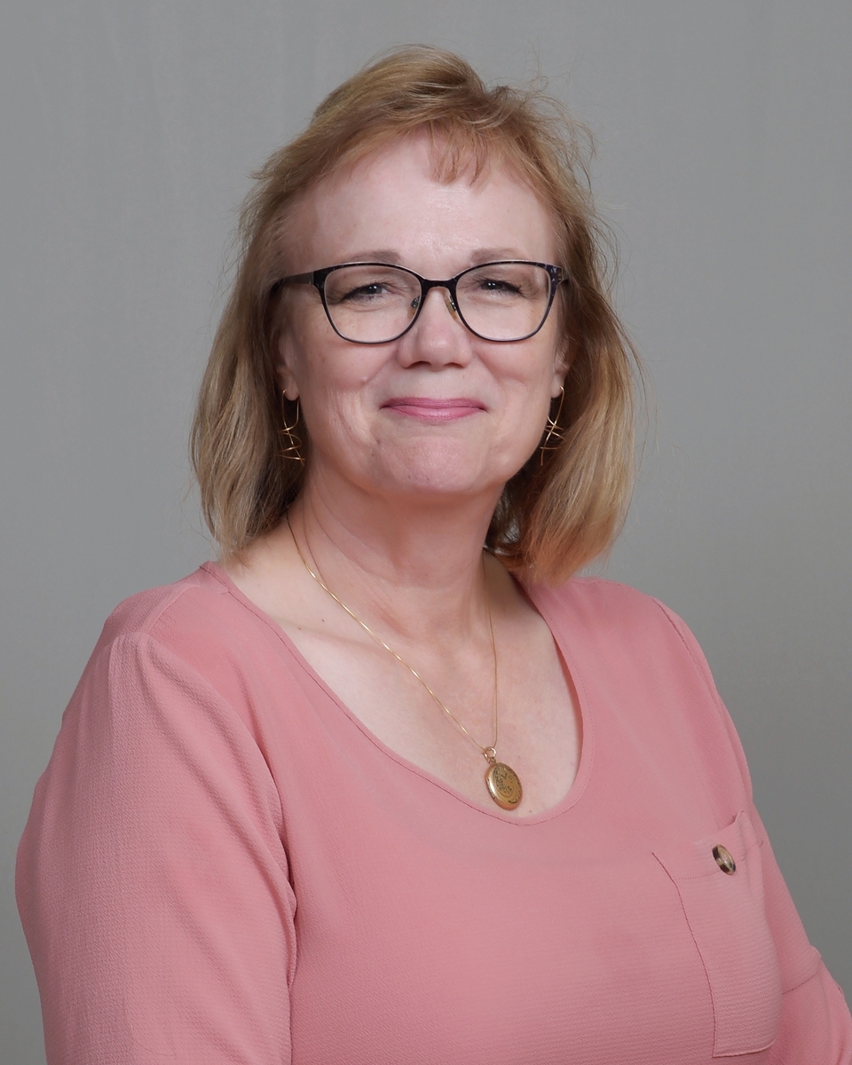 Lisa is smiling at the camera. She has reddish-blonde shoulder length hair, glasses and is wearing a coral pink blouse. She has on a gold chain necklace with a gold locket.