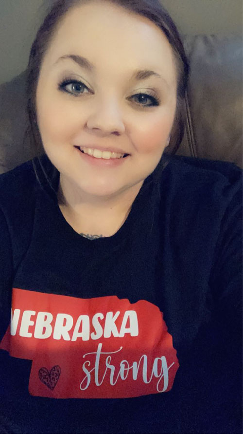 Young woman with long dark hair, dark eyes, and a black shirt with the words Nebraska Strong and a heart on a red image of the state of Nebraska