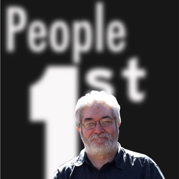 Un hombre con el pelo y la barba canosos de pie delante de un gran texto que dice «People 1st» (Las personas primero).