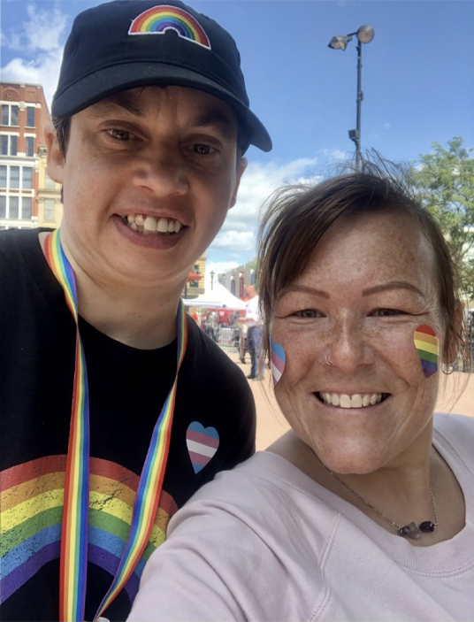 Dos mujeres con logotipos arco iris sonríen mientras miran al frente. Una lleva una gorra y una camiseta negra, la otra una camisa lila.