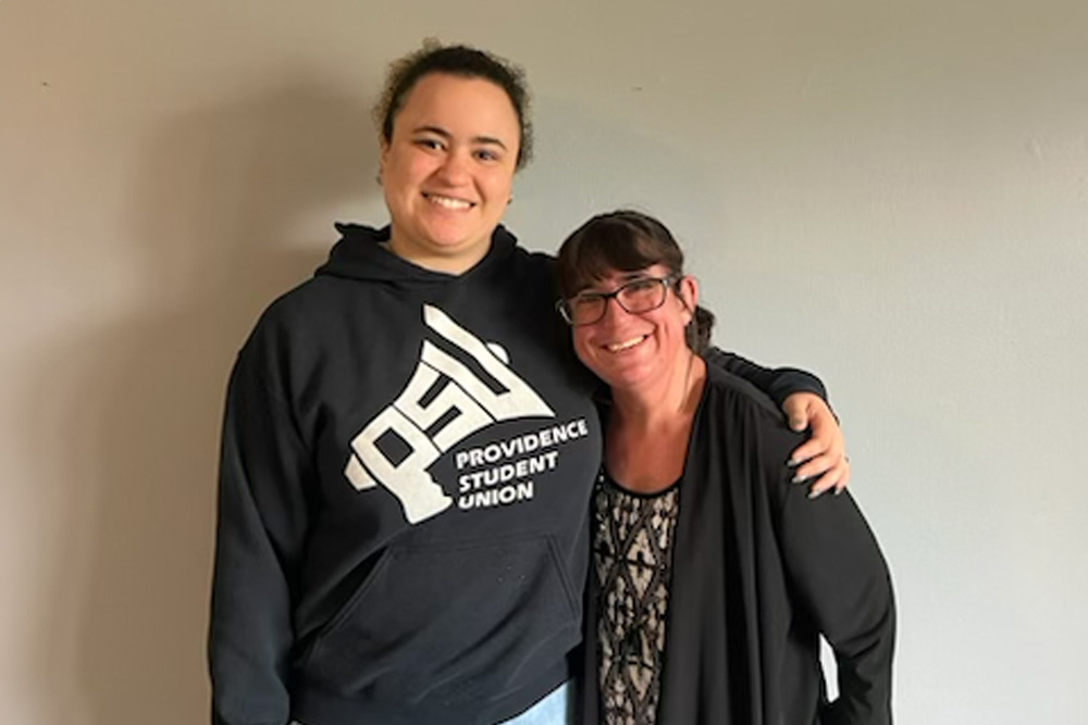 Roselin y Becky están una al lado de la otra, sonriendo a la cámara. Roselin está a la izquierda y lleva vaqueros y una sudadera negra con capucha que dice Providence Student Union con PSU en forma de megáfono. Roselin tiene el cabello oscuro que parece recogido hacia atrás. Becky se encuentra a la derecha. Lleva unos vaqueros negros y un jersey, con un top blanco y negro con rombos y tiras diagonales. Tiene el cabello oscuro recogido en una coleta y lleva gafas de montura negra. Roselin tiene su brazo izquierdo alrededor del hombro de Becky.