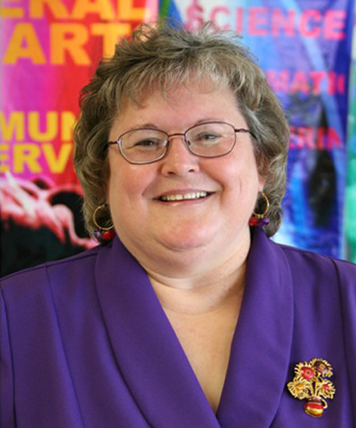 Woman with blonde-brown hair, glasses, ear rings, purple top with big smile.