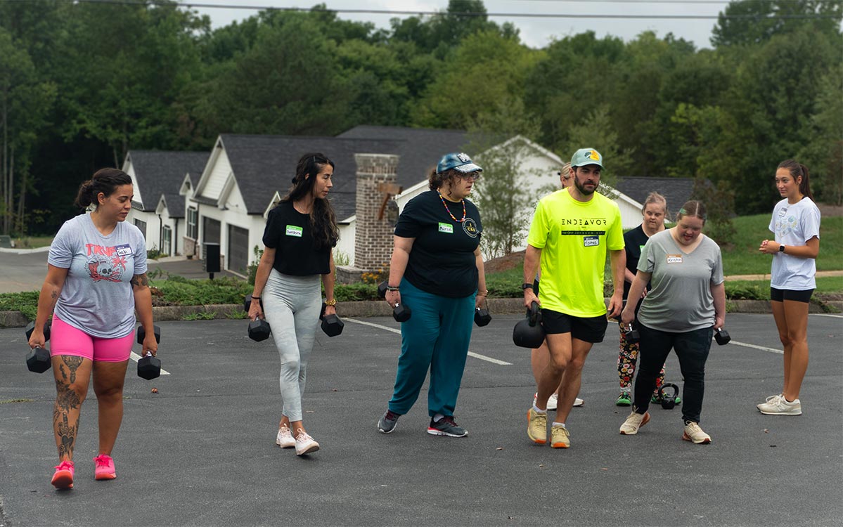 Seis personas caminan juntas al aire libre mientras transportan pesas en un ejercicio de Farmer Carry. El grupo incluye a participantes de I CAN y miembros del gimnasio que entrenan juntos durante una sesión de Buddy Workout.