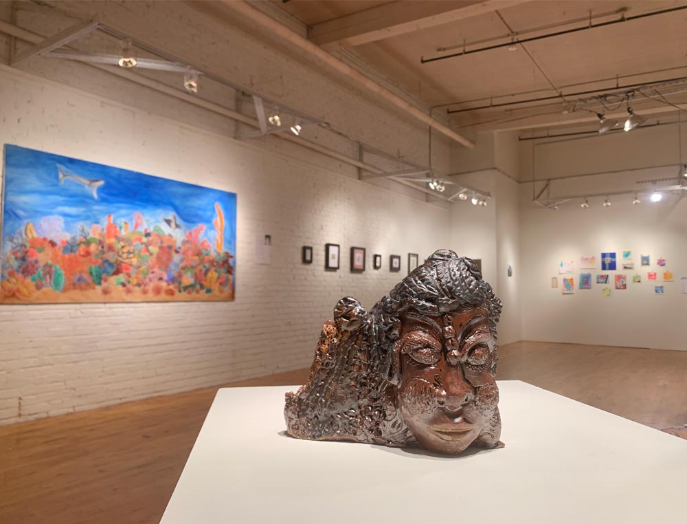 A ceramic piece featuring a face sits on a small display stand with paintings in the background during an art show.