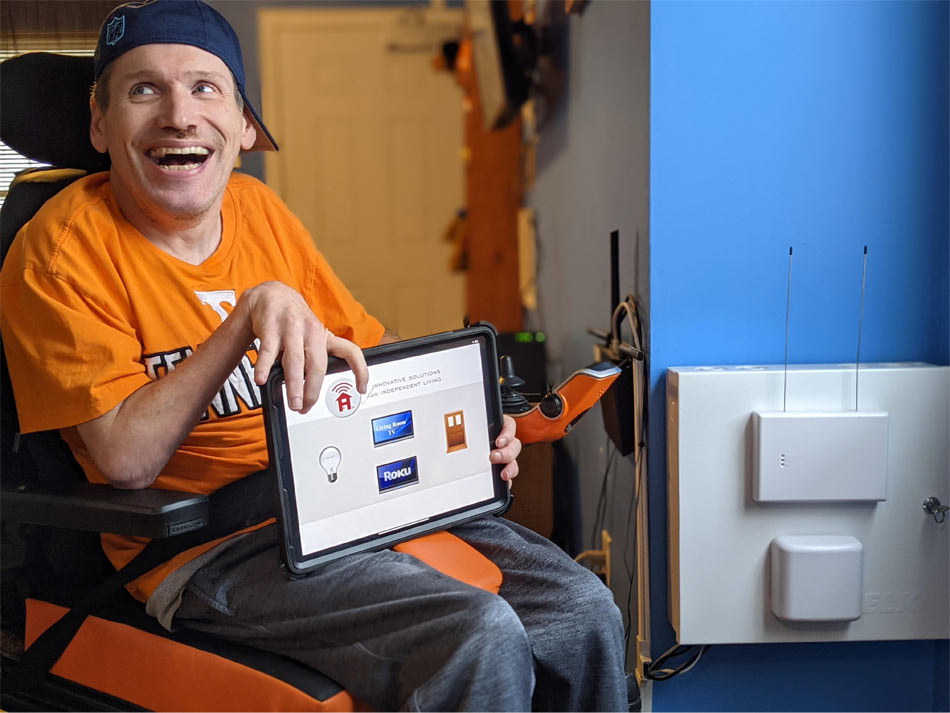 Un hombre está sonriendo y mirando hacia arriba y a su derecha. Él está sosteniendo un i-pad en sus manos. La pantalla del i-pad está mirando hacia la cámara e ilustra la aplicación que utiliza para abrir las puertas de su casa, junto con otras aplicaciones como Roku. Él está sentado en su silla de ruedas, que está adornada con asientos naranjas. Lleva un sombrero de estilo béisbol oscuro hacia atrás, y una camiseta naranja de los voluntarios de Tennessee con una gran T blanca en la parte superior y Tennessee en el pecho. Él está usando pantalones deportivos grises.