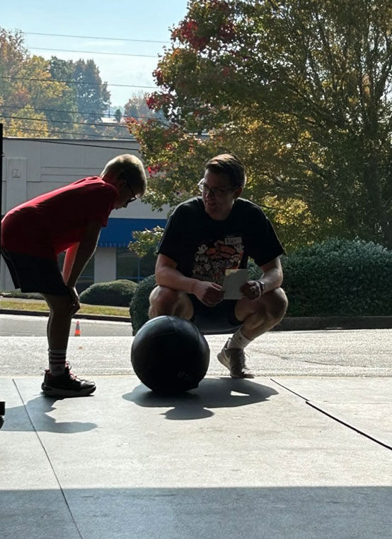 Alex, a community gym member, encourages Landon, an I CAN participant, during ball slams at a Buddy Workout. Alex cheers while Landon takes a break between sets.