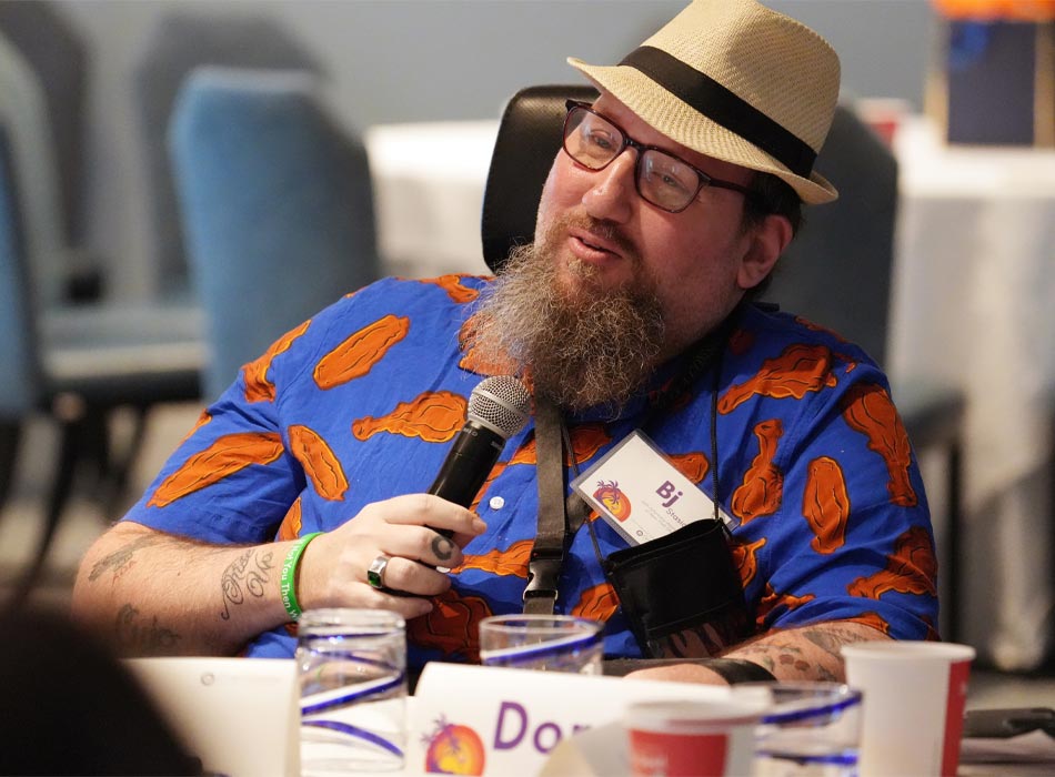 A bearded man wearing a straw fedora hat and a bright blue shirt with orange designs holds a microphone.