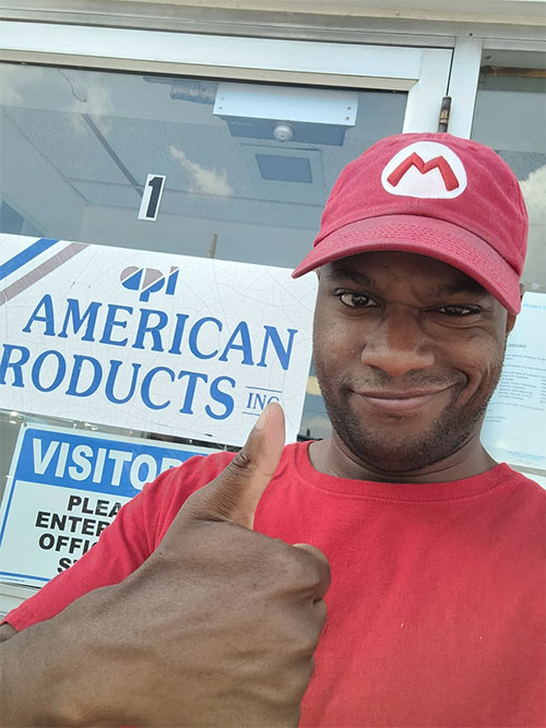 Cody muestra un gesto de aprobación con el pulgar delante de una puerta con el rótulo que dice "American Products, Inc". Lleva una camiseta roja y una gorra de béisbol roja con una "M".