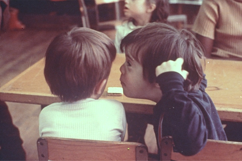 A child with Down syndrome whispers in the ear of a classmate.