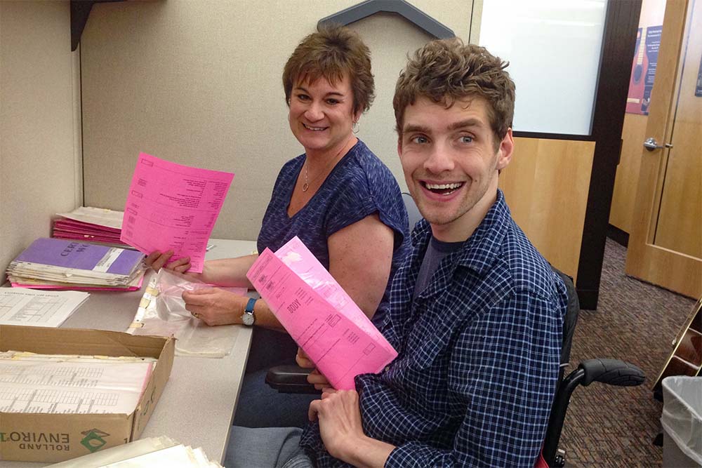 Two individuals with light skin are seated at a desk in an office-like setting, working with pink paper forms. The person on the left, a woman with short brown hair, is wearing a blue patterned top and smiling at the camera. The person on the right, a young man with curly brown hair uses a wheelchair, is wearing a blue plaid shirt and smiling broadly. The desk has stacks of papers and a cardboard box containing files, and the background includes beige walls and a wooden door.