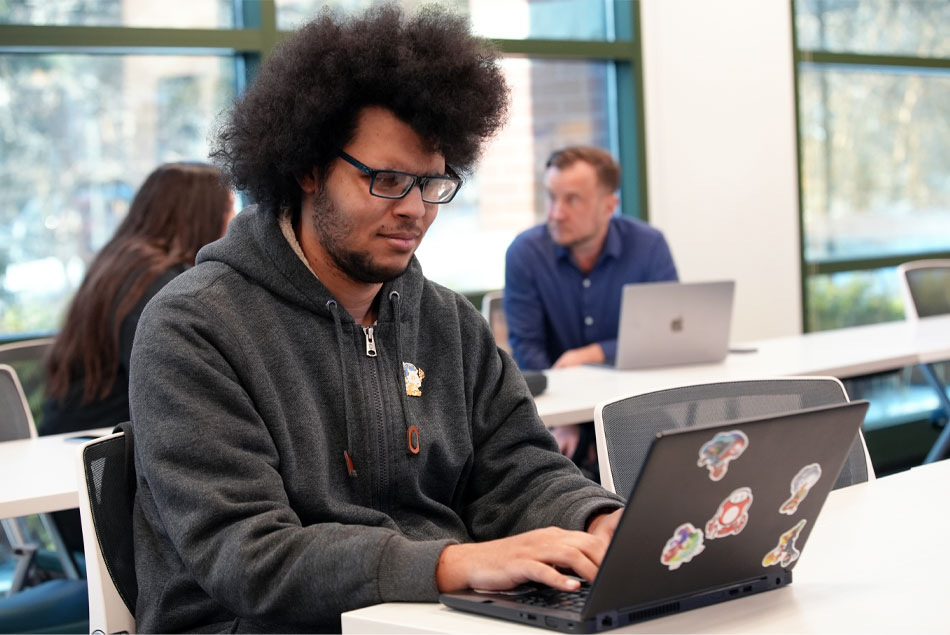 Un hombre sentado a una mesa, con un peinado afro natural, gafas de montura oscura y una sudadera con capucha, mira con seriedad una computadora portátil mientras está sentado frente a otras dos personas, cuyos rostros están borrosos.
