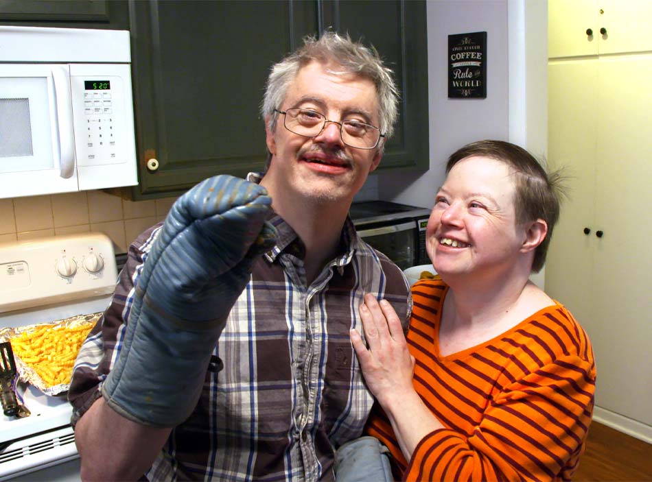 A man with gray hair and glasses smiles as a woman with short dark hair places her hand on his shoulder.