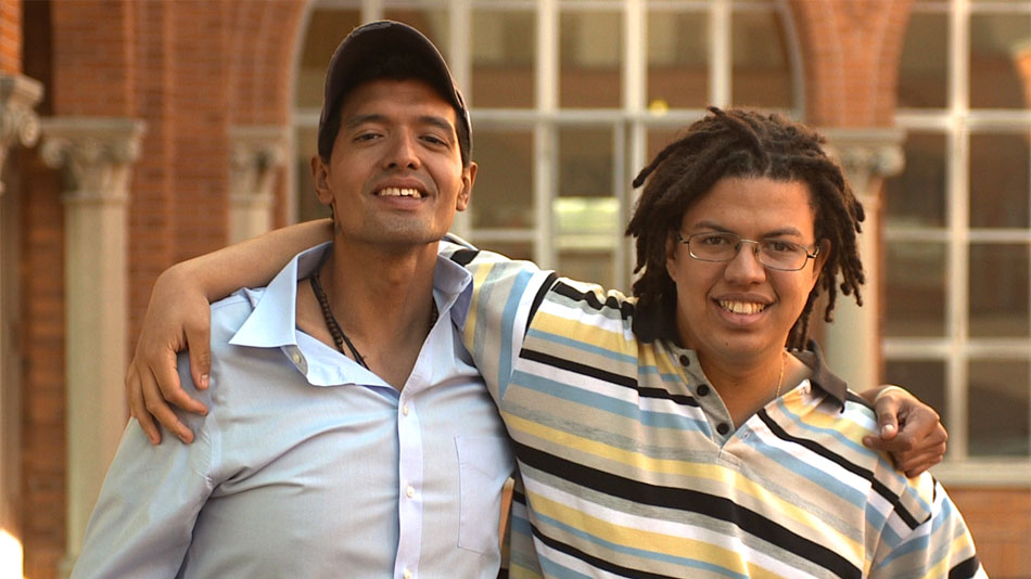 Two young men smile, with their arms around each other.
