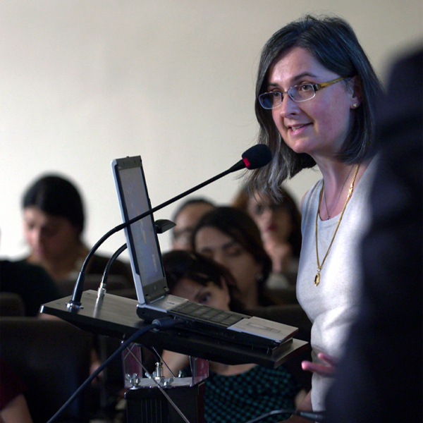 Mujer con cabello oscuro hasta los hombros y gafas hablando por un micrófono en un aula.
