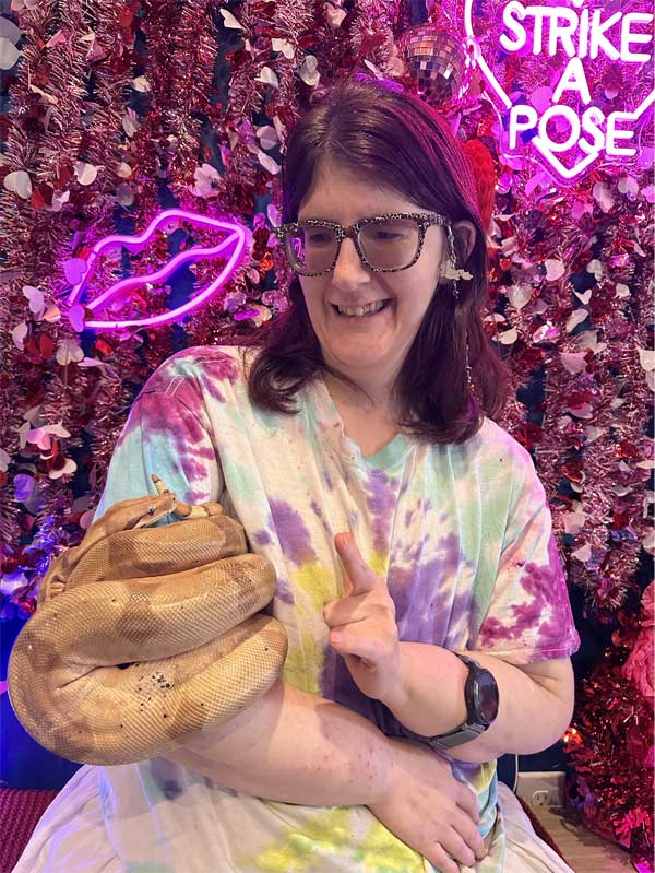 A woman with speckled glasses and brown hair holds her pet snake and signs “I love you” as she poses in front of a pink background, wearing a tie-dyed shirt.
