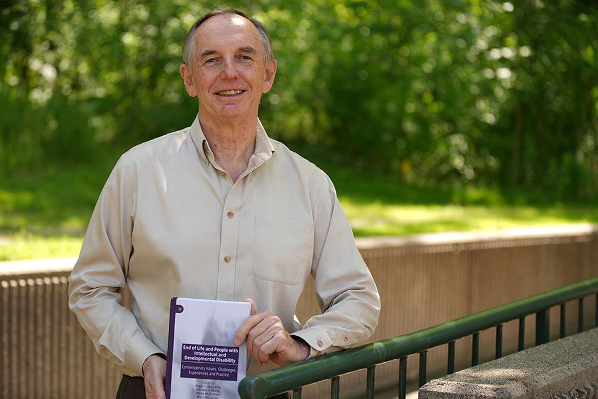 Roger is standing outside. There are green trees in the background. He has short graying hair, is wearing a tan long sleeve button down shirt. He is holding a book in front of him that is facing the camera. The book he has authored titled: End of life and people with intellectual and developmental disabilities.