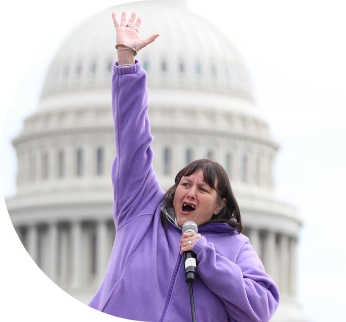 A woman with dark hair wearing a lilac jacket speaks excitedly while holding a microphone.