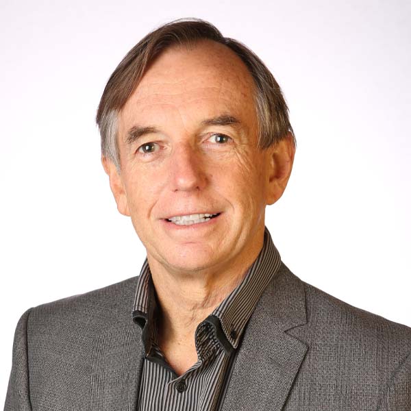 studio photograph of a man with short dark hair that is slightly graying, smiling and looking at the camera. He is wearing a dark suit and dark shirt.