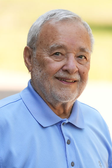An older man with light skin tone and short white hair is smiling. He is wearing a light blue polo shirt. The background is softly blurred, featuring greenery and a neutral tone.