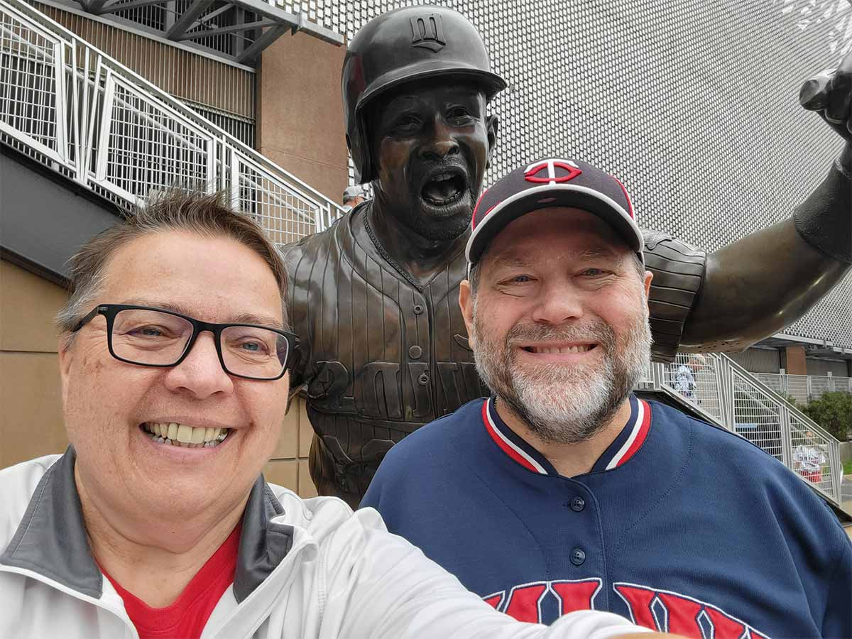 Dos personas delante de la estatua de Kirby Pucket. Chet es la mujer de la izquierda. Ella sonríe, tiene el cabello corto castaño y blanco, lleva gafas y viste una camiseta roja con un jersey blanco y gris. Jimmy es el hombre de la derecha. Sonríe, tiene una barba corta marrón y blanca, y lleva una gorra de béisbol azul, roja y blanca de los Minnesota Twins y un jersey azul marino.