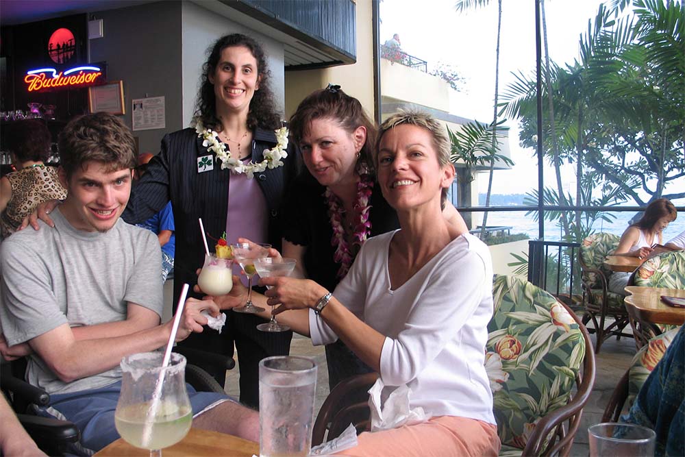 Four people are enjoying happy hour at an indoor/outdoor restaurant ocean side. Renzo is setting and holding a fruity drink with a pineapple and cherry on top, two people stand to the right, and with Hawaiian leis, one holds a martini. The fourth woman is across from Renzo, holding a fancy glass of water and supporting his glass. They are smiling and looking at the camera.